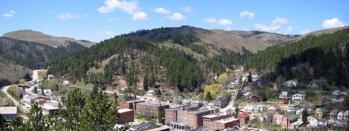 A panoramic view of a small town nestled in green hills, with buildings and trees under a blue sky with scattered clouds.