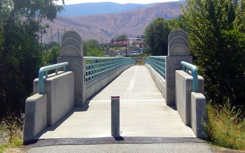 A concrete bridge with blue railings spans a pathway, surrounded by trees and hills in the background.