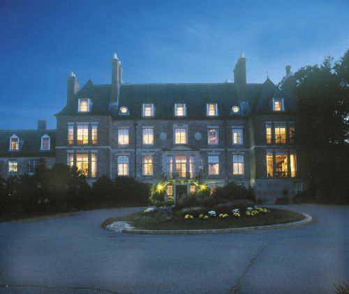 A grand stone mansion illuminated at dusk, surrounded by landscaped gardens and a circular driveway.