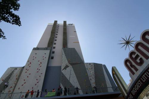 A tall climbing wall on a building with climbers, under a clear blue sky, near a sign that reads "Reno."