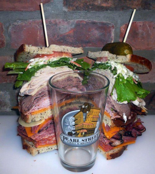 A towering sandwich stacked with meats, cheese, and greens, accompanied by a pint glass and a pickle on top.