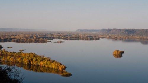 A serene view of a river surrounded by autumn foliage and distant hills under a clear sky.