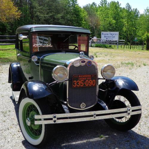 A vintage green car with a black hood and white wheels parked outdoors, surrounded by trees and a sign in the background.