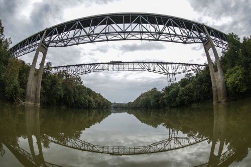 A wide river reflects two large, parallel bridges above, surrounded by lush greenery and a cloudy sky.
