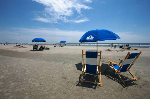 Two beach chairs under blue umbrellas on a sandy beach, with people enjoying the sun and ocean in the background.
