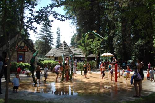 Children and adults enjoy a splash pad in a park, surrounded by trees and a play area with water features.