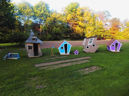 A row of colorful, whimsical miniature houses on a grassy field, surrounded by trees and a clear blue sky.