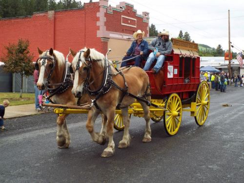 A horse-drawn wagon with two horses pulls a cart, while two men sit on it during a parade in a small town.