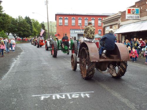A parade of vintage tractors approaches the finish line, with spectators lining the street in a small town.