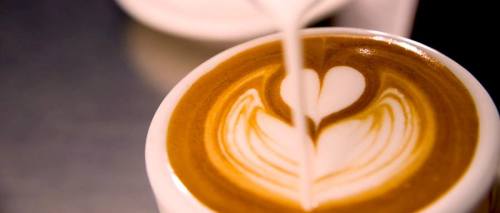 A close-up of a latte with a heart-shaped foam design being poured into a white cup.