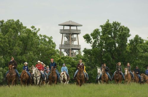A group of people on horseback riding through a grassy area, with a lookout tower in the background.