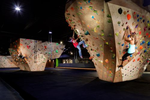Two climbers ascend colorful bouldering walls in a modern indoor climbing gym, with soft mats below for safety.
