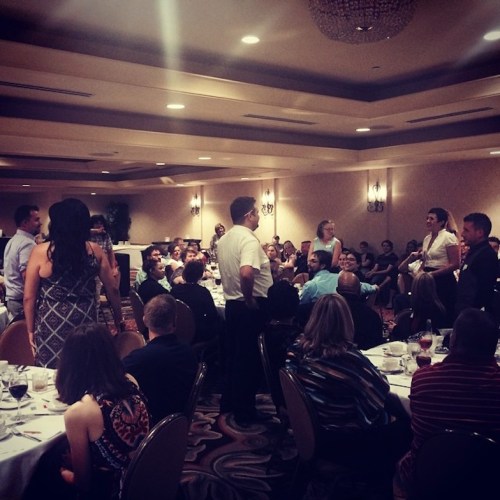 A large group of people seated at tables in a banquet hall, with some standing and engaging in conversation.