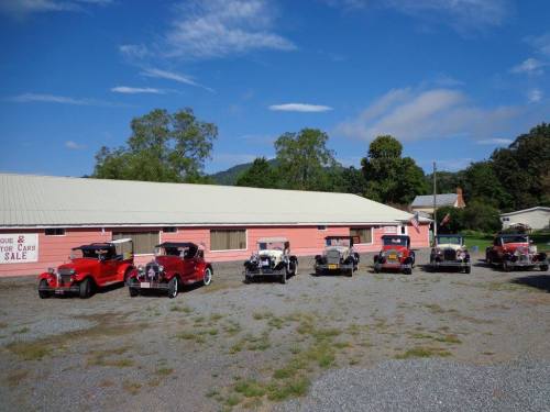 A row of vintage cars parked in front of a pink building with a sign for "Cars for Sale" under a clear blue sky.