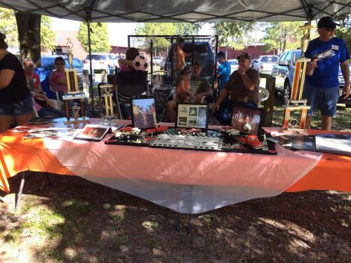 A table displays trophies and memorabilia under a tent, with people seated nearby in a park setting.
