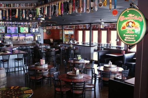 Interior of a brewery with wooden tables, chairs, and a bar, decorated with hanging beer taps and a sign.