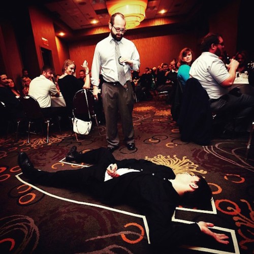 A man in a suit lies on the floor outlined in white, while another man stands nearby, looking surprised in a crowded room.