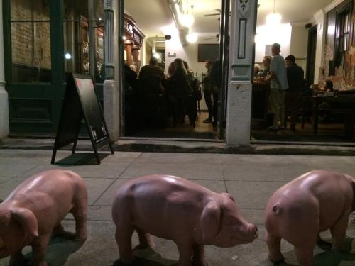 Three pink pig sculptures are positioned on the sidewalk in front of a bustling restaurant with patrons inside.