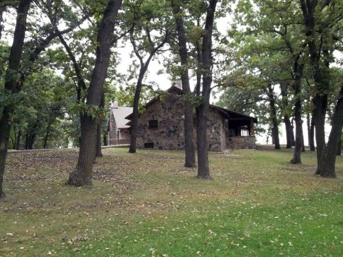 A rustic stone house surrounded by tall trees and grassy land, set in a serene outdoor environment.