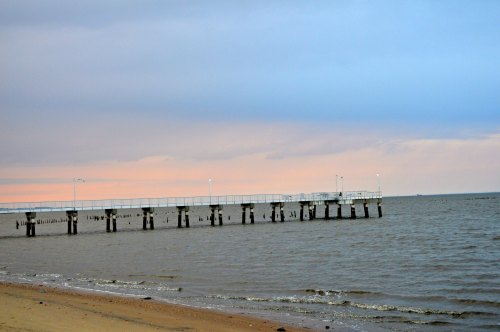 A wooden pier extends over calm waters, with a sandy beach in the foreground and a pastel sky at dusk.