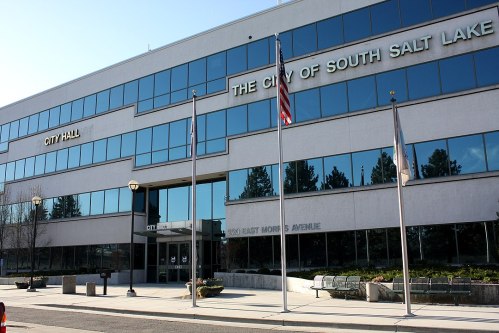 City Hall building in South Salt Lake, featuring large windows and flags in front, with a clear blue sky above.
