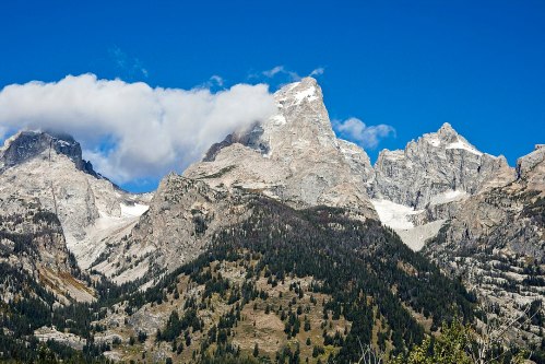 Majestic mountain range under a clear blue sky, with rocky peaks and patches of snow amidst green forests.