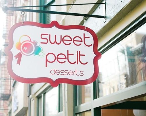 Sign for "Sweet Petit Desserts" hanging outside a shop, featuring colorful graphics and a playful design.