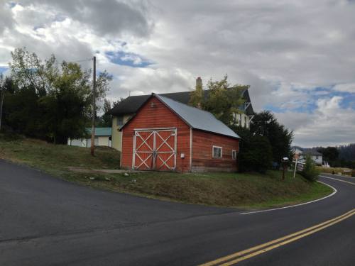 A red barn with a metal roof at a road intersection, surrounded by trees and houses under a cloudy sky.