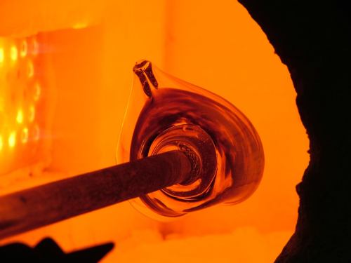 A close-up of molten glass being shaped in a furnace, glowing orange with a glassblowing pipe inserted.