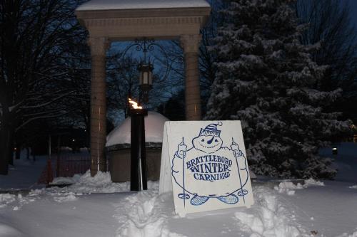 A sign for the Brattleboro Winter Carnival stands in the snow, with a torch burning nearby and trees in the background.