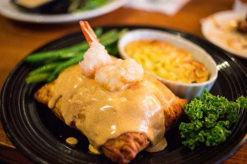 A plate featuring a shrimp-topped pastry with creamy sauce, green beans, and a small dish of baked macaroni.