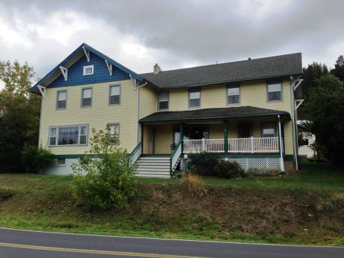 A large yellow house with blue accents, featuring a porch and surrounded by greenery, under a cloudy sky.