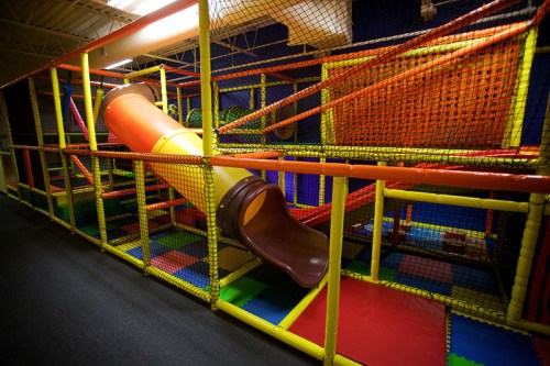 Colorful indoor play structure with a large slide and climbing areas, surrounded by safety netting and soft flooring.