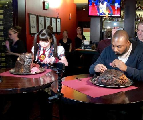 A girl in a school uniform and a man sit at a table, each with large portions of meat on platters, preparing to eat.