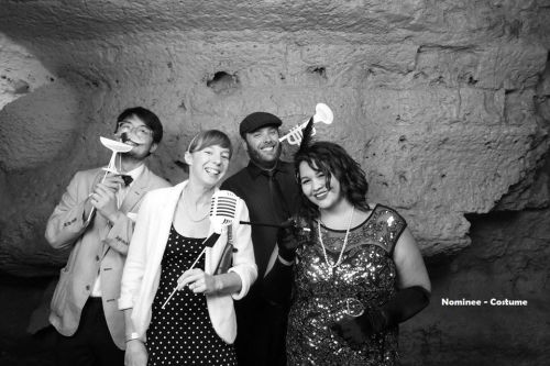 A group of four friends in vintage attire, posing playfully with props in a cave-like setting.