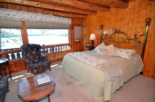 Cozy wooden bedroom with a large bed, floral armchair, and lake view through large windows.