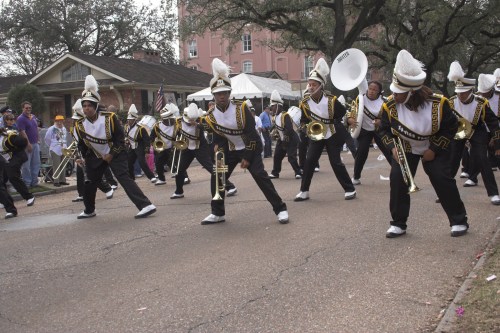 A marching band in black and gold uniforms performs on a street, playing instruments and moving in unison.