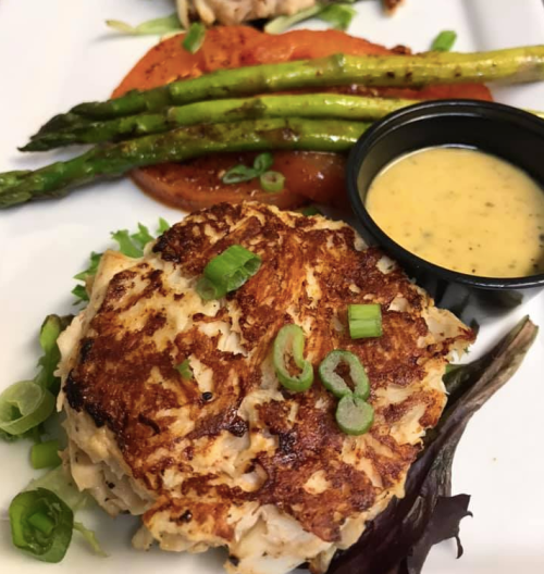 A plate featuring a crab cake topped with green onions, asparagus, roasted tomato, and a small cup of dipping sauce.