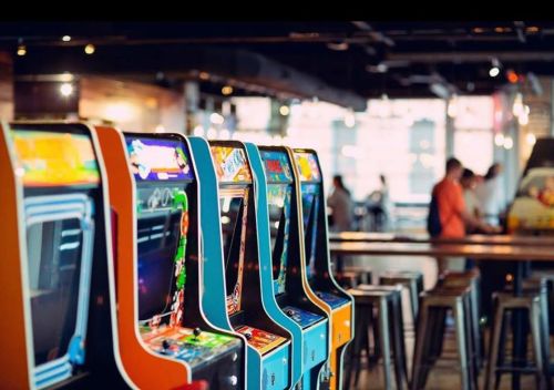 A row of colorful arcade game machines in a modern gaming venue, with people in the background.