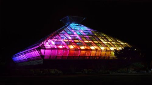 A brightly lit, colorful glass pyramid structure at night, showcasing vibrant hues of purple, blue, and orange.