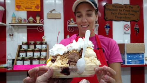 A smiling ice cream shop employee holds a large sundae topped with whipped cream, cherries, and chocolate.