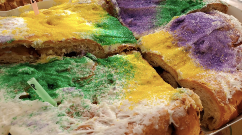 A close-up of a colorful king cake, decorated with yellow, green, and purple sugar, cut into pieces.