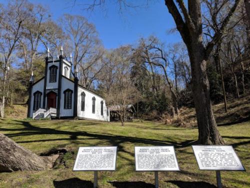 A small white church with a red door, surrounded by trees and historical markers on a sunny day.