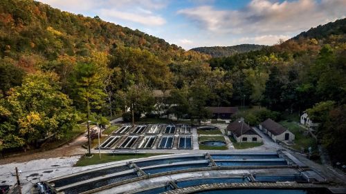 Aerial view of a fish hatchery surrounded by lush green hills and trees under a partly cloudy sky.