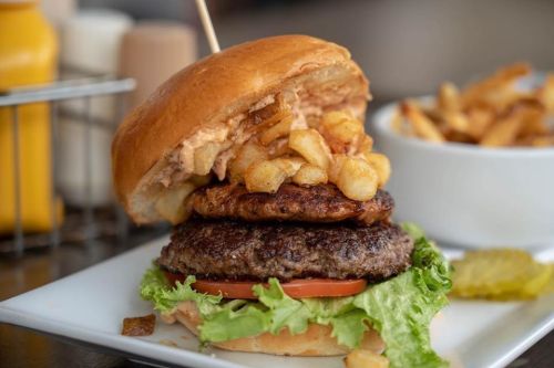 A stacked burger with two patties, lettuce, tomato, and crispy fries on top, served with a side of fries.
