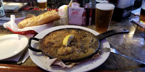 A skillet of baked seafood with a lemon slice, served with a side of bread and a glass of beer.