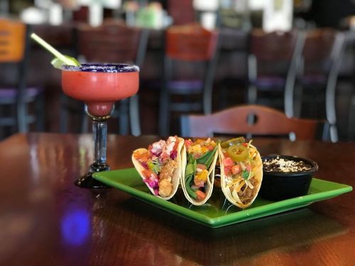 Three tacos filled with toppings on a green plate, accompanied by a small bowl of black beans and a colorful margarita.