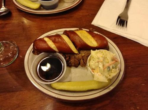 A plate with two pretzel sausages, coleslaw, a pickle, and a small cup of dipping sauce on a wooden table.