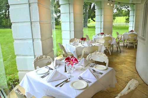 Elegant dining area with white tables, floral centerpieces, and large windows overlooking a green landscape.