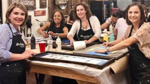 Four women in aprons work on art projects at a crafting table, smiling and engaged in their creative activities.